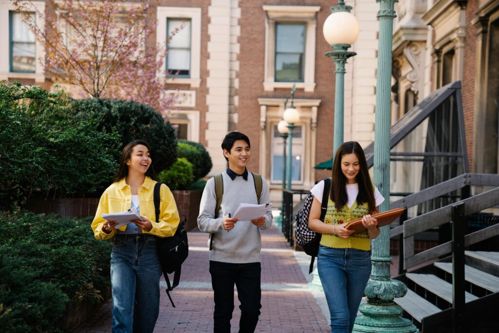 Three young adults walking and talking on a sunny campus day, holding notebooks and backpacks.
